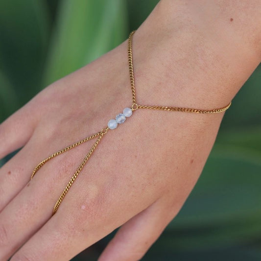 A delicate gold hand chain with three small aquamarine beads, shown worn on a person's hand against a blurred green background.