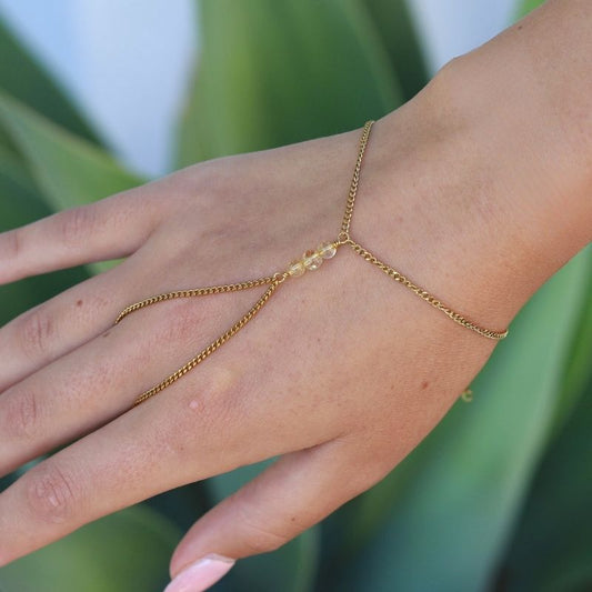 A delicate gold hand chain with a small cluster of citrine beads at the center, shown worn on a person's hand against a green leaf background.