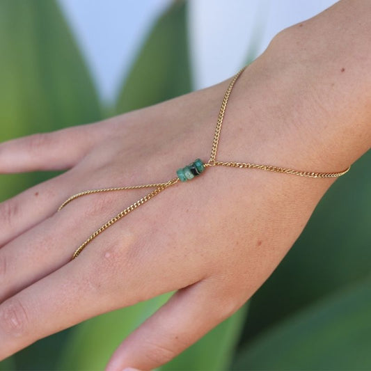 A delicate gold hand chain with a cluster of small emerald stones worn on a person's hand and wrist, shown in a close-up shot against a blurred green background.