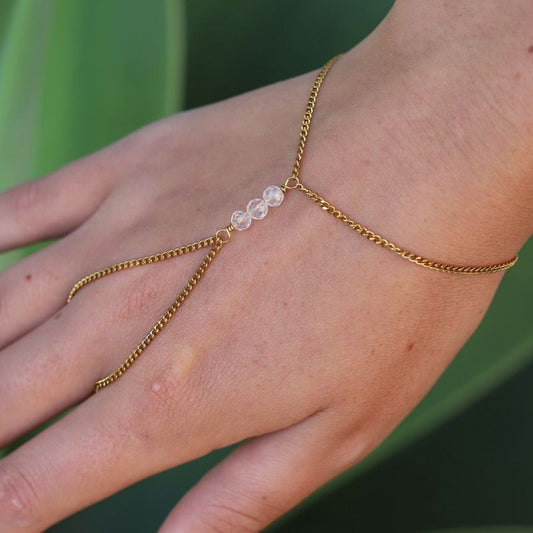 A delicate gold hand chain with three small quartz crystal beads, shown worn on a person's wrist and hand against a blurred green background.