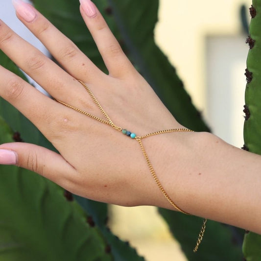 A gold hand chain with three small turquoise-blue beads at the center, shown worn on a person's hand with green leaves in the background.