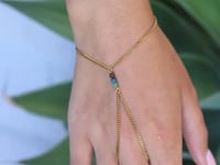 A slowly moving hand wearing a gold hand chain with three small turquoise-blue beads at the center, shown worn on against a blurred background with soft green leaves.