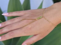 A slowly moving hand wearing a delicate gold hand chain with three small peridot beads at the center, shown worn against a blurred green background.