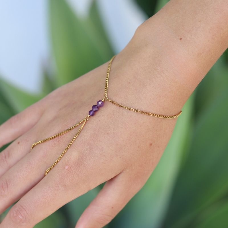 A delicate gold hand chain with three small amethyst beads at the center, shown worn on a person's hand against a soft green blurred background.
