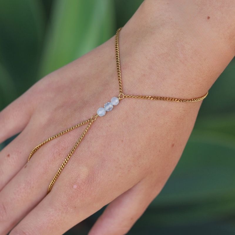 A delicate gold hand chain with three small aquamarine beads, shown worn on a person's hand against a blurred green background.