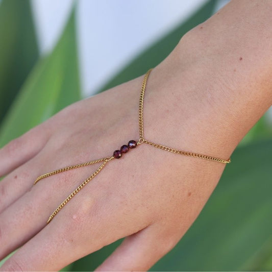 A delicate gold hand chain with three small garnet beads at the center, shown worn on a person's hand against a soft green blurred background.