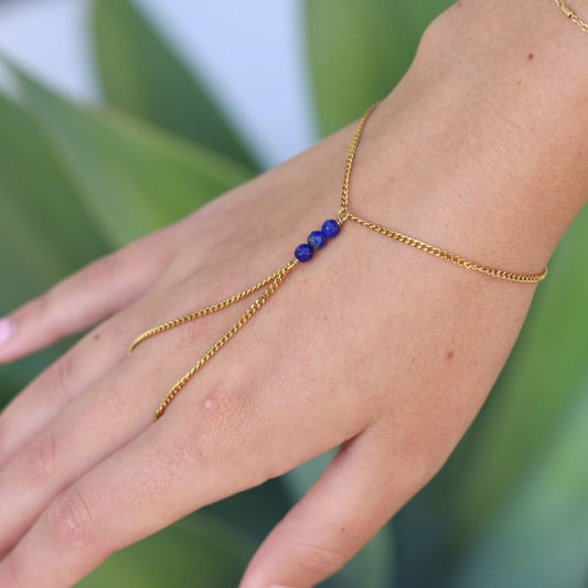 A delicate gold hand chain with three blue lapis lazuli beads at the center, shown worn on a person's hand against a soft green leaf background.