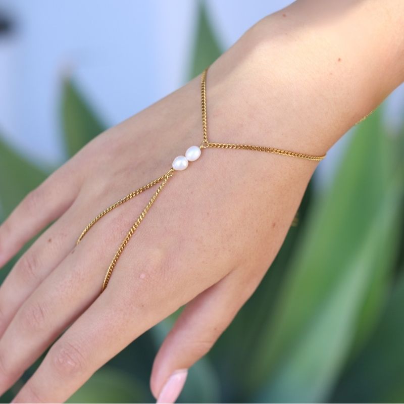A delicate gold hand chain with two small white pearls, shown worn on a person's wrist and hand against a soft-focus green background.
