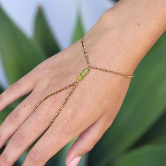 A delicate gold hand chain with three small peridot beads at the center, shown worn on a person's hand against a blurred green background.