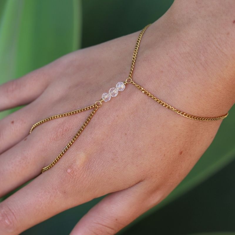 A delicate gold hand chain with three small quartz crystal beads, shown worn on a person's wrist and hand against a blurred green background.