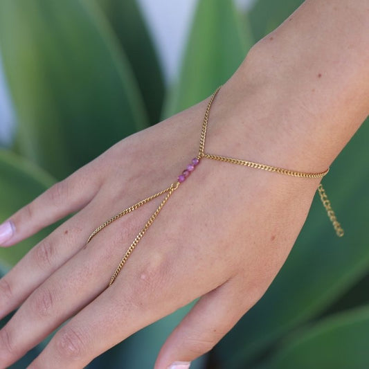 A delicate gold hand chain with three small ruby beads, shown worn on a person's wrist and hand against a blurred green background.