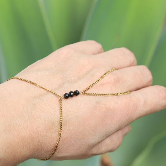 A delicate gold hand chain with three small black tourmaline beads, shown worn on a person's hand against a soft green blurred background.