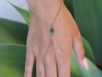 A slowly moving hand wearing a delicate gold hand chain with a cluster of small emerald stones, shown worn in a close-up shot against a blurred green background.