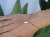 A slowly moving hand wearing a  delicate gold hand chain with two small white pearls, shown worn against a soft-focus green background.