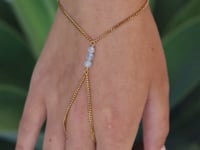 A slowly moving hand wearing a  delicate gold hand chain with three small aquamarine beads, shown worn against a blurred green background.