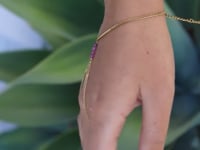 A slowly moving hand wearing a delicate gold hand chain with three small ruby beads, shown worn against a blurred green background.