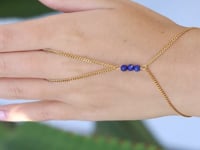 A slowly moving hand wearing a  delicate gold hand chain with three blue lapis lazuli beads at the center, shown worn against a soft green leaf background.