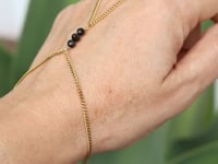 A slowly moving hand wearing a delicate gold hand chain with three small black tourmaline beads, shown worn against a soft green blurred background.