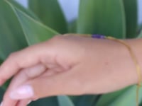 A slowly moving hand wearing a delicate gold hand chain with three small amethyst beads at the center, shown worn against a soft green blurred background.